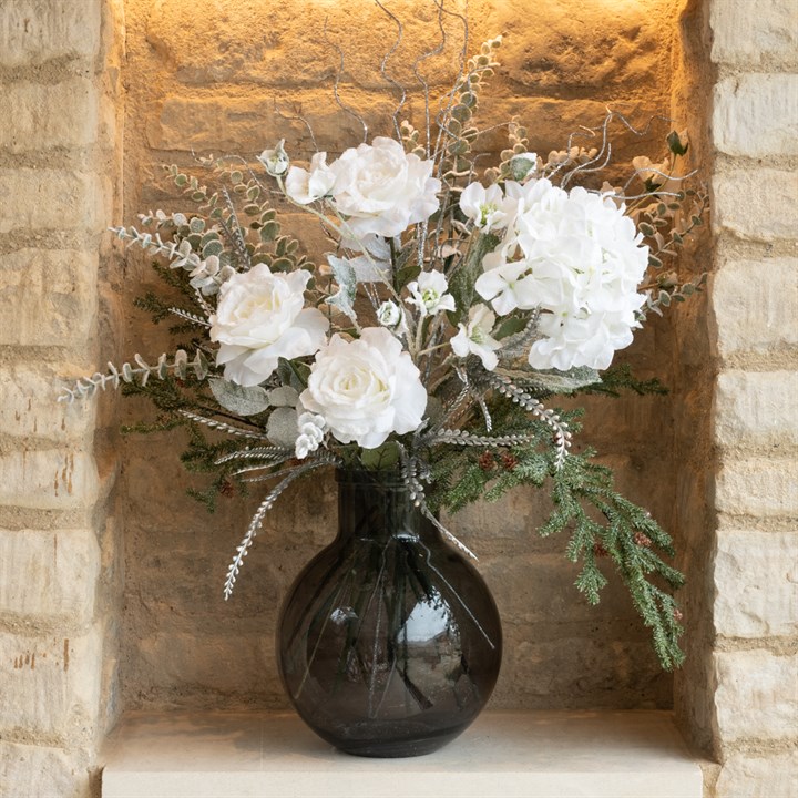 white faux flowers and foliage arranged in a glass vase in an alcove