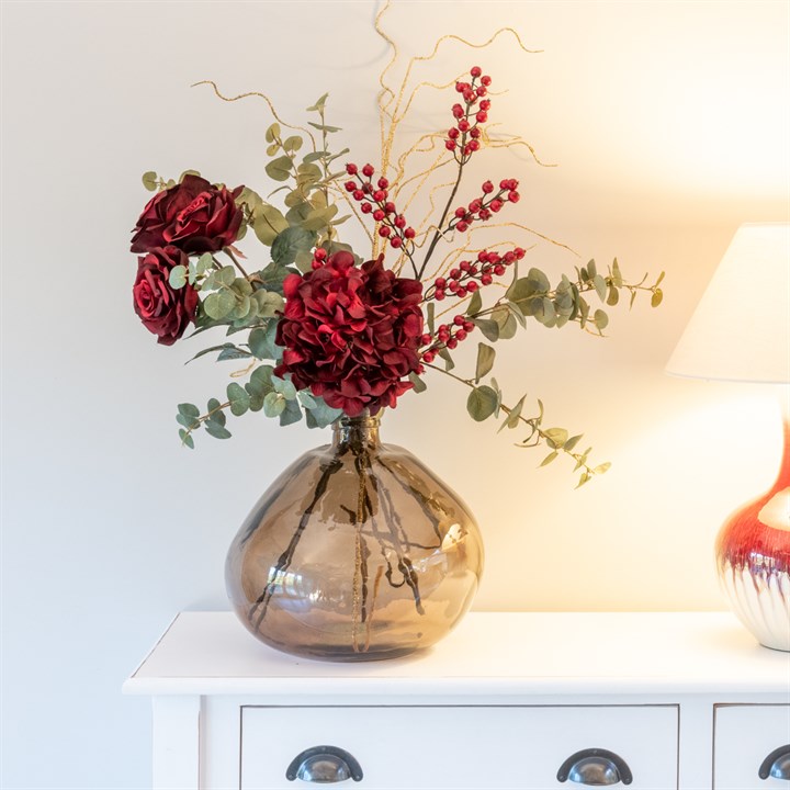 red faux flowers and berries with eucalytpus in brown glass vase on dresser