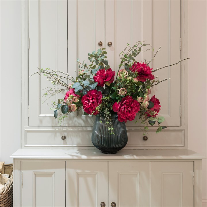 red faux flowers and green foliage arranged in glass vase on a dresser