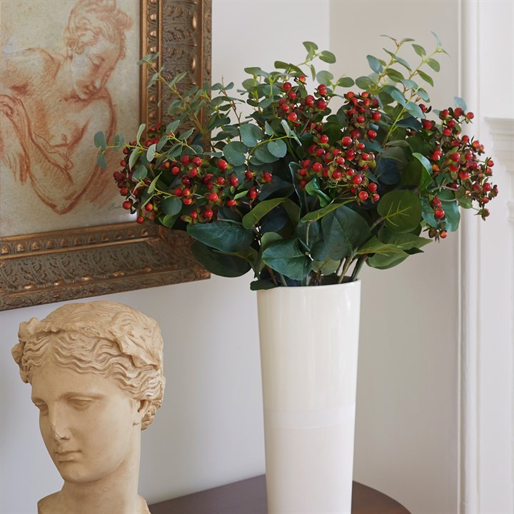green artificial foliage and berries arranged in tall white vase with painting and bust sculpture