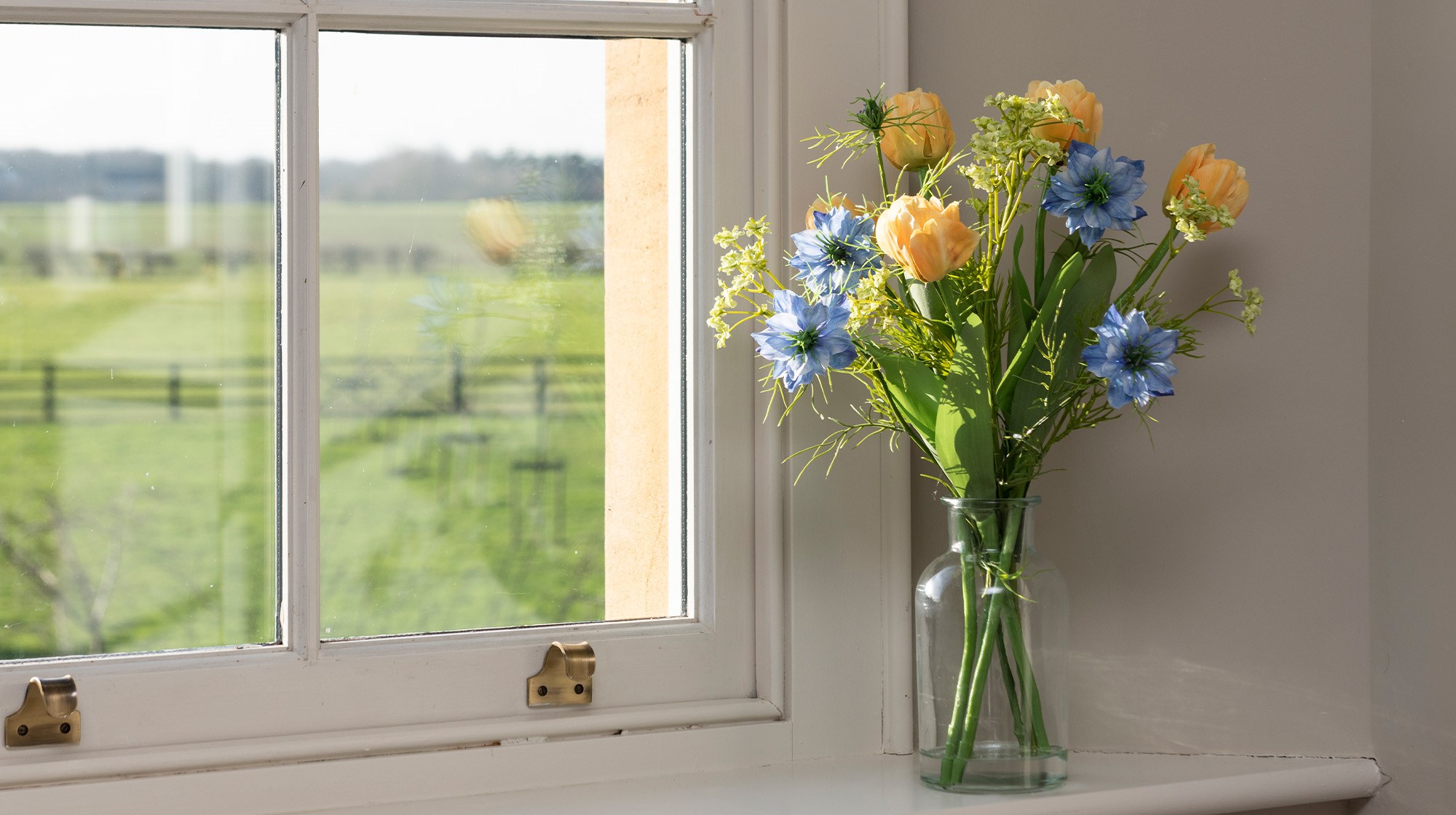A faux tulip and nigella arrangement in a small clear vase, set on a windowsill