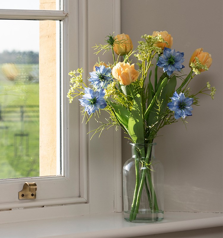 A realistic artificial tulip and nigella arrangement in a small clear vase, set on a windowsill